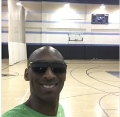 a man wearing sunglasses and smiling for the camera in a gym with basketball hoop behind him