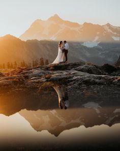 a bride and groom standing on top of a mountain with their reflection in the water