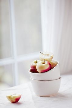 two white bowls filled with apples sitting on top of a table next to a window