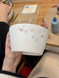 a person is holding up a white paper bowl with flowers on it and paint supplies nearby