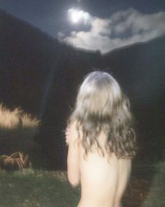 a naked woman sitting on top of a lush green field under a moon filled sky