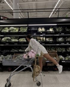 a woman pushing a shopping cart through a grocery store filled with vegetables and flowers,