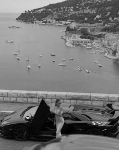 a woman standing next to a black sports car on the side of a road with boats in the water