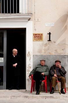 two men sitting on red chairs in front of an open door and another man standing next to them