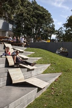 people sitting on benches in the grass near some stairs and trees, while others walk by