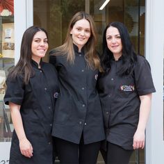 three women standing in front of a pastry shop wearing black uniforms and smiling at the camera