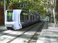 an electric train traveling down the tracks next to a tree lined sidewalk with people walking on it