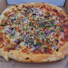 a pizza with onions, cheese and herbs in a cardboard box on a table top