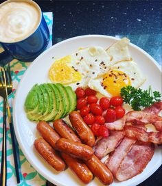 a white plate topped with eggs, bacon, tomatoes and avocado next to a cup of coffee