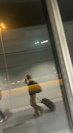 two people are walking with their luggage through an airport terminal at night, and one person is using his cell phone