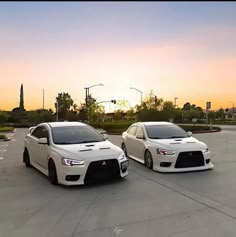 two white cars parked next to each other in a parking lot with the sun setting behind them