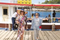 two women in hats and sunglasses standing on a dock near the water with their arms outstretched