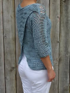 a woman standing in front of a wooden fence wearing a crocheted sweater and white pants