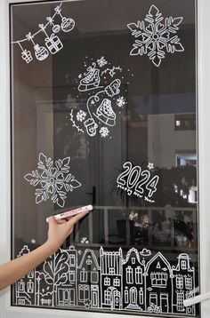 a person writing on a glass window with white snowflakes and houses drawn on it