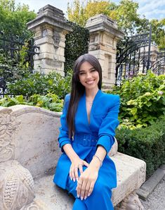 a woman sitting on a stone bench in front of some bushes and trees, wearing a blue suit