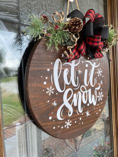 a wooden sign that says let it snow hanging on the front door with pine cones and evergreens