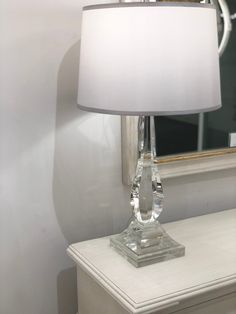 a clear glass table lamp sitting on top of a white dresser next to a mirror