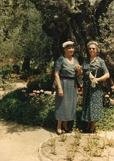 two older women standing next to each other in front of a large tree and flowers