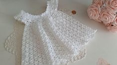 a crocheted baby dress next to pink roses on a white table with doily