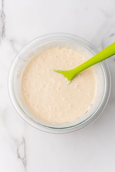 a glass bowl filled with batter on top of a white counter next to a green spoon