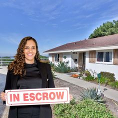 a woman standing in front of a house holding a sign that says in escrow