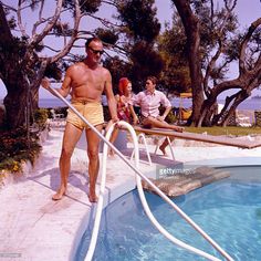 a man with no shirt and yellow shorts standing next to a swimming pool while two other people sit in the background