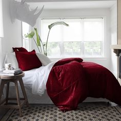 a bed with red sheets and pillows in a room next to a window, near a small table