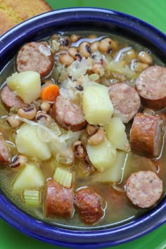 a blue bowl filled with sausage and potatoes next to a piece of bread on a green plate