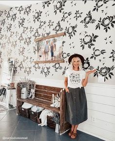 a woman standing next to a wooden bench in front of a wall with flowers on it