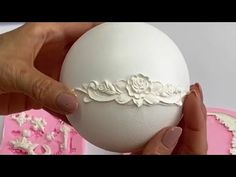 a woman is holding an egg decorated with white flowers and lace on the top, in front of a pink cake