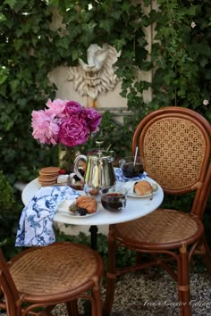 two chairs and a table with food on it in front of some bushes, one has pink flowers