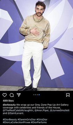 a man standing in front of a purple wall wearing white pants and a turtle neck sweater