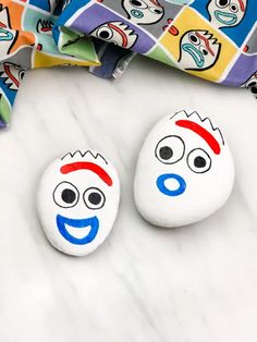 two painted rocks sitting on top of a white counter next to a colorful cloth with cartoon faces