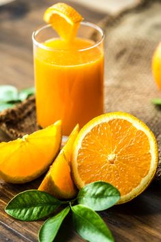 an orange juice in a glass next to sliced oranges and leaves on a wooden table