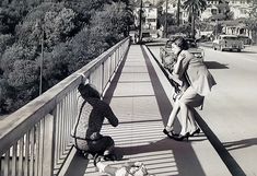 two women are standing on a bridge looking at the street below them and one is holding a teddy bear