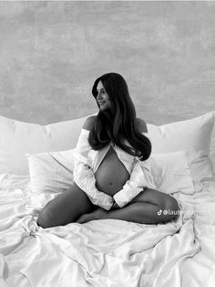 a pregnant woman sitting on top of a bed with white sheets and pillows in front of her