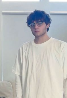 a young man wearing glasses standing in front of a white wall