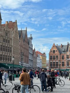 many people are walking and riding bikes in the city square with old buildings on either side