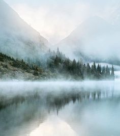 a lake surrounded by mountains with fog in the air and trees on it's sides