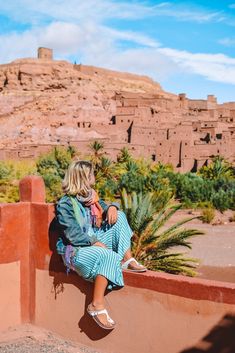 a woman sitting on top of a red wall next to palm trees and a mountain