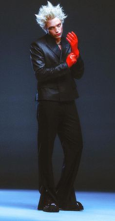 a male in a black suit and red gloves is standing with his arms folded up