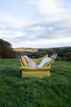 a woman laying on top of a yellow chair in the grass with her feet up