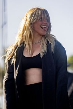 a woman with blonde hair wearing a black jacket and cropped top smiles at the camera