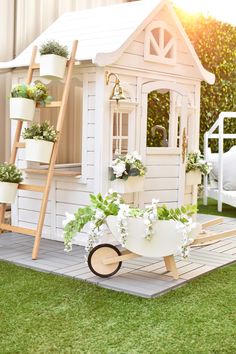 a small white house with potted plants on the roof and wheelbarrow in front