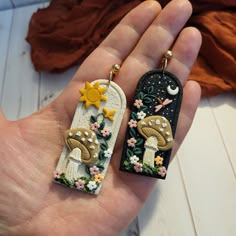 two handmade earrings with mushrooms and flowers on them are being held by someone's hand