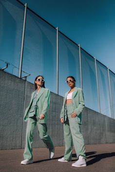 two women standing next to each other in front of a fence wearing green suits and sunglasses