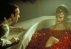 a man and woman sitting in a bathtub with red petals on the floor, looking at each other