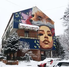 a large building with a painting on it's side in the snow next to parked cars