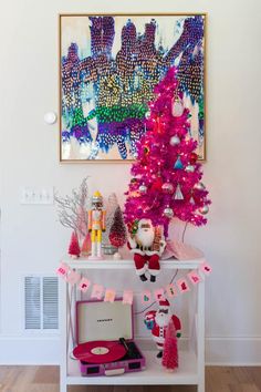 a pink christmas tree in front of a painting and other decorations on a white table