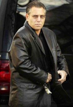 a man in a black suit and sunglasses is walking towards the camera with his hand on his hip